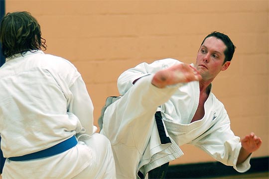 A black belt man is doing a hook kick toward a color belt student durning free sparring at an All-School workout.