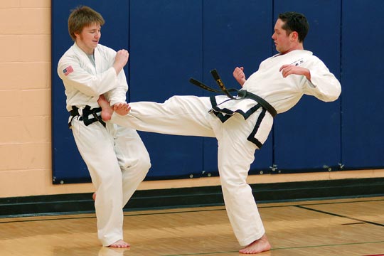 Two black belt men free-spar during an All-School workout. The man on the right lands a twisting kick to the belt.