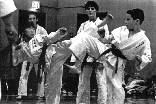 A girl and boy spar during a tournament. The girl is executing a high roundhouse kick.