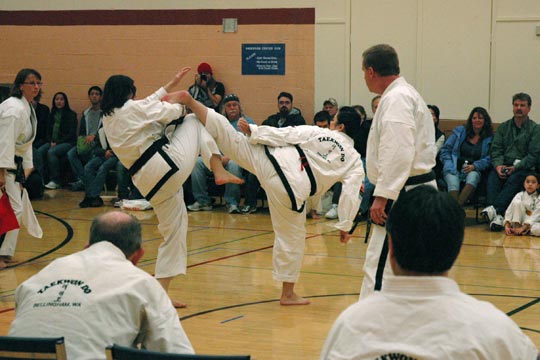 Two black belt women spar at a tournament. One is attempting a kick toward the other's head.