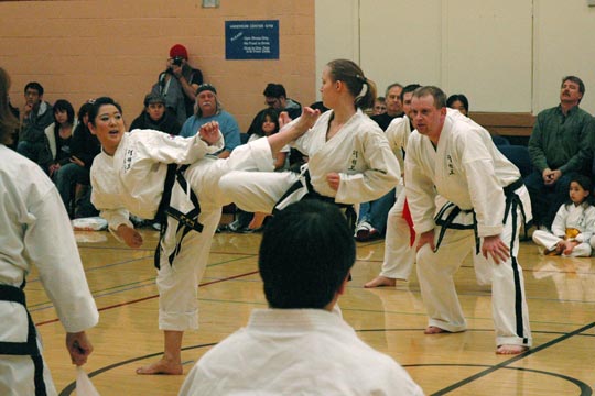 Two black belt women spar at a tournament. One is attempting a roundhouse kick toward the other's head.