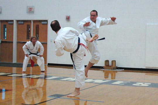 Two blue belt men spar at a tournament. The man on the ground is starting a kick toward the one in the air. The man in the air appears to have his lead knee high enough to block his opponents potential kick.