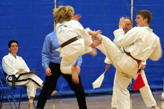Two brown belts spar at a tournament. One is in the air blocking the other's kick.