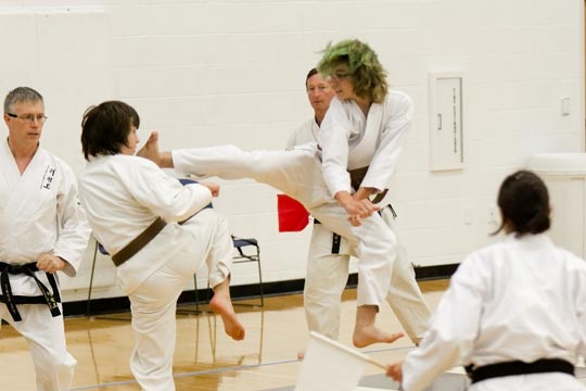 A brown belt with green tinged hair is landing a jumping kick to the face of his opponent.