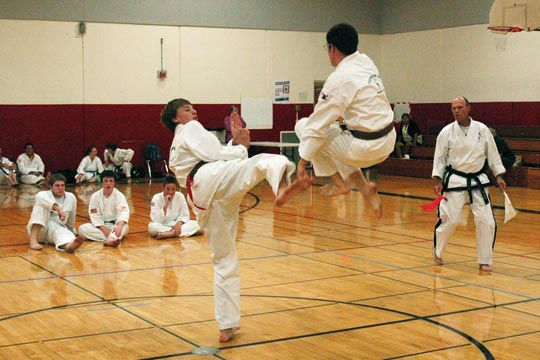 Two brown belt men spar at a tournament. The one in the air has both feet tucked up higher than the belt of his standing opponent.