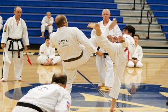 Two brown belt men spar at a tournament. Both are doing head level kicks toward the other.