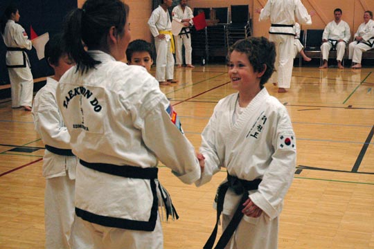 A black belt woman with her back to the camera shakes the hand of a smiling brown belt boy.