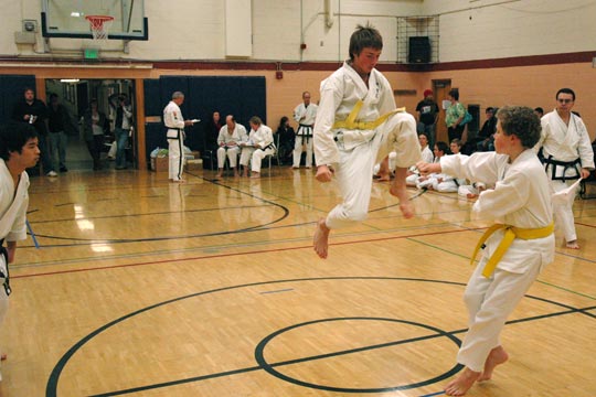 Two yellow belt boys spar at a tournament. One is attempting a jumping kick while the other blocks and moves back.