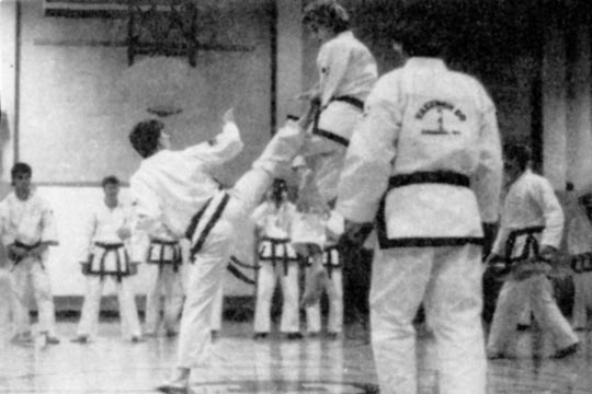 Two men are tournament sparring. The man jumping is being kicked by the man standing.