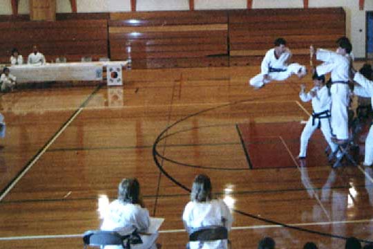 A man does a jumping twisting kick to a board held over another man's head.