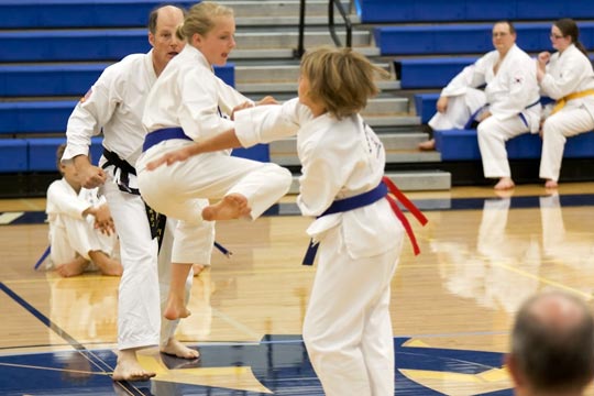 Two blue belt girls spar at a tournament. The one on the ground is extending a knifehand weapon. The one in the air is executing a roundhouse kick.