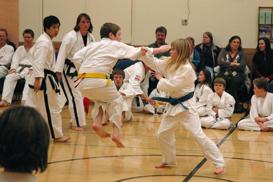 A yellow belt boy does a jumping knifehand strike outward toward a blue belt girl during a tournament match. The girl is blocking the attack.