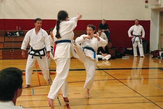 Two blue women spar at a tournament. The one facing the camera is landing a front kick on the other.