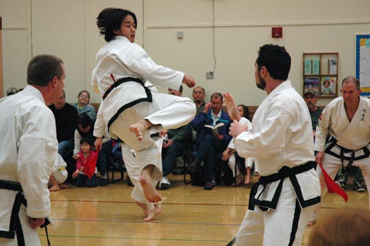 Two black belt men spar at a tournament. One is attempting a jumping kick toward the other.