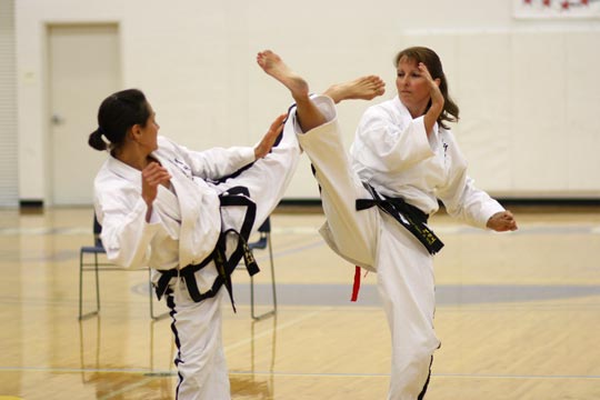 Two black belt women spar at a tournament. One is attempting a twisting kick to the head while the other attempts a roundhouse kick to the head.