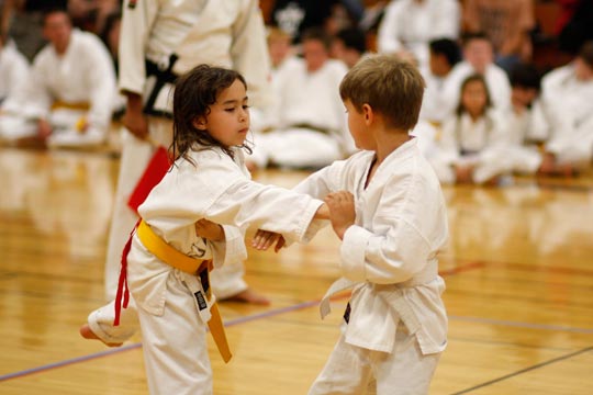 A white belt boy and yellow belt girl spar during a tournament. The girl is throwing a punch which her apponent is attempting to block.
