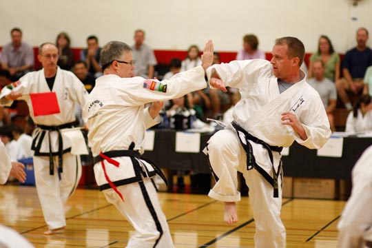 Two black belt men spar at a tournament. The one wearing glasses appears to be only moments away from getting kicked in the stomach.