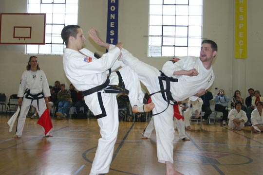 Two men spar during a tournament. One is extending a high kick toward the other's head.