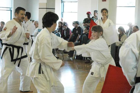 Two junior white belts spare at a tournament. Judges and competitors are all smiling.