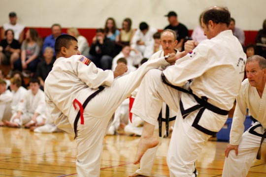 Two black belt men spar at a tournament. The shorter man is trying to land a side kick which the taller man is blocking.