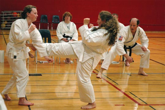 Two white belt women spar at a tournament. The woman on the right is landing a ball-of-the-foot front kick while the woman on the left tries to block it.